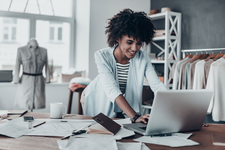 A woman looks at a laptop with papers and clothing on racks and on a mannequin behind her. Photo by iStock.