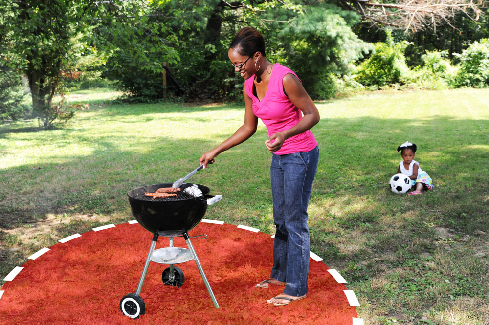 A woman cooks meat on a charcoal grill outside. There is circle with a 3-foot radius drawn around the grill. A young girl plays with a soccer ball well outside of the circle. Image by U.S. Fire.