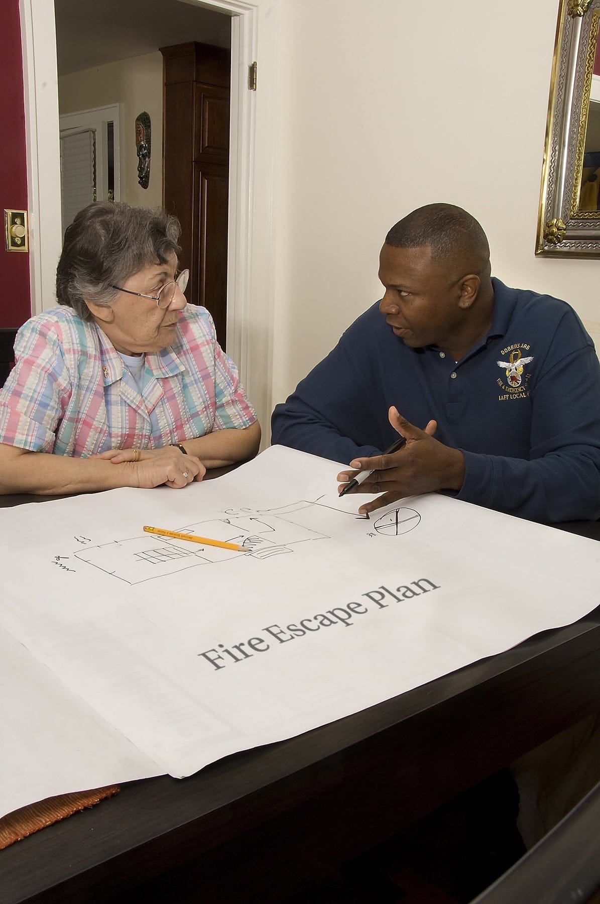 An older woman listens to a younger man discuss a fire escape plan drawing that is located in front of them both.