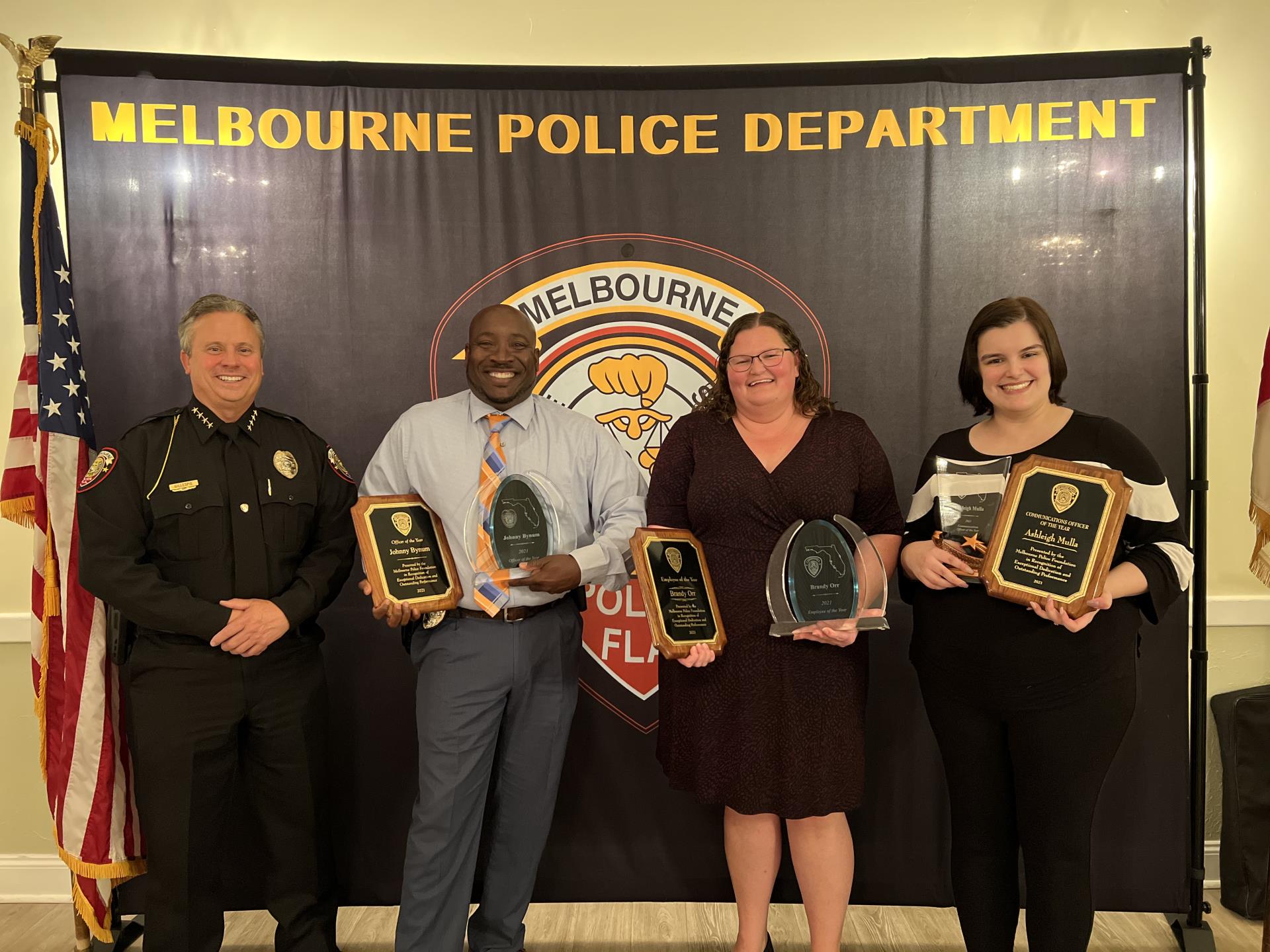 Chief Gillespie with Detective Bynum, Brandi Orr and Ashleigh Mulla, who are holding awards and standing in front of the Melbourne Police Department backdrop.