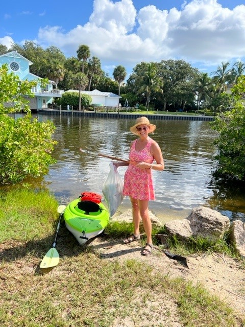 A kayaker stands next to her kayak and a bag of trash at Ballard Park with the Eau Gallie River behind her.